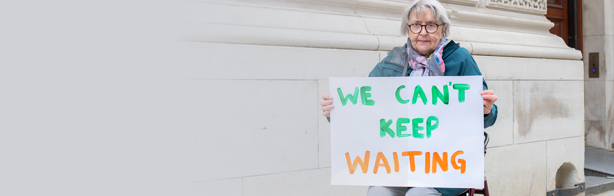 Older woman with sign reading 'We can't keep waiting'