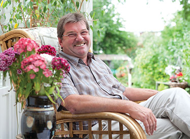 A man sat in a wooden chair in his conservatory surrounded by plants